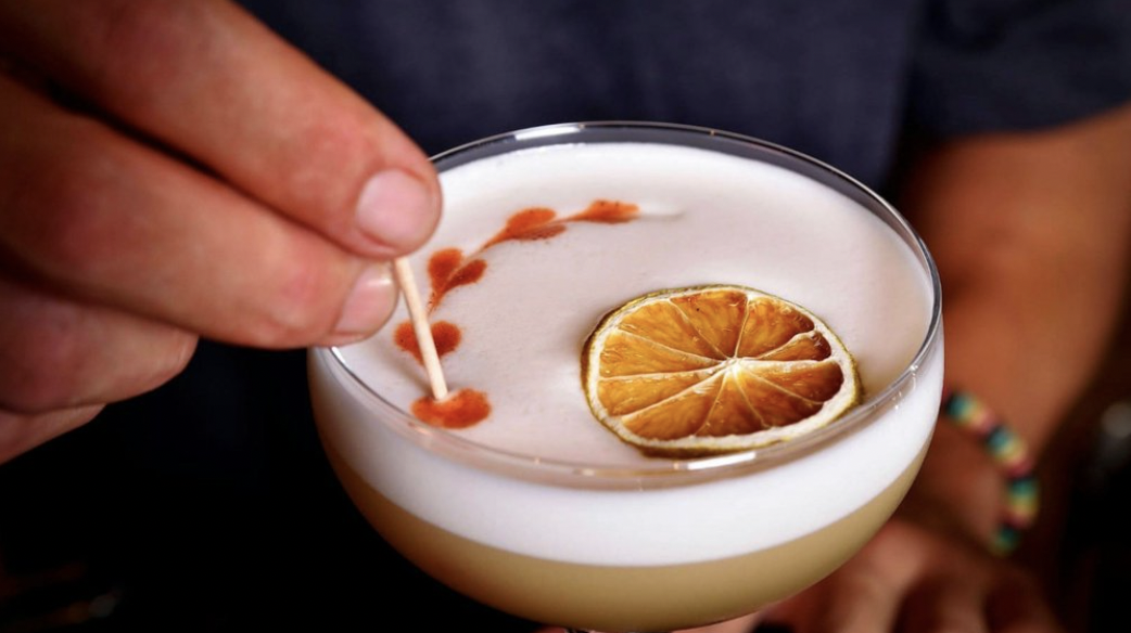 A bartender is using a toothpick to draw a design in a Pisco Sour cocktail