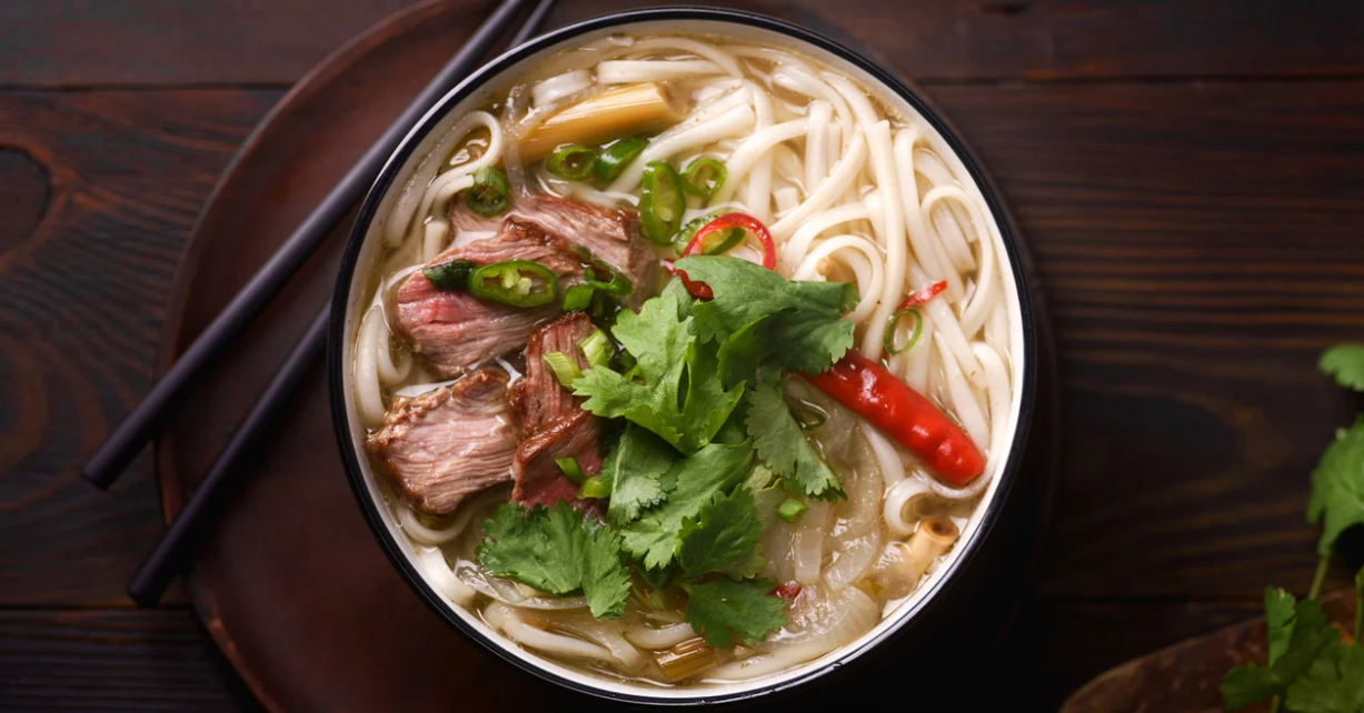 (Bovenaanzicht van een kom Vietnamese pho met dikke rijstnoedels, plakjes rundvlees, verse koriander, rode en groene chilipepers en uien in een heldere bouillon.)