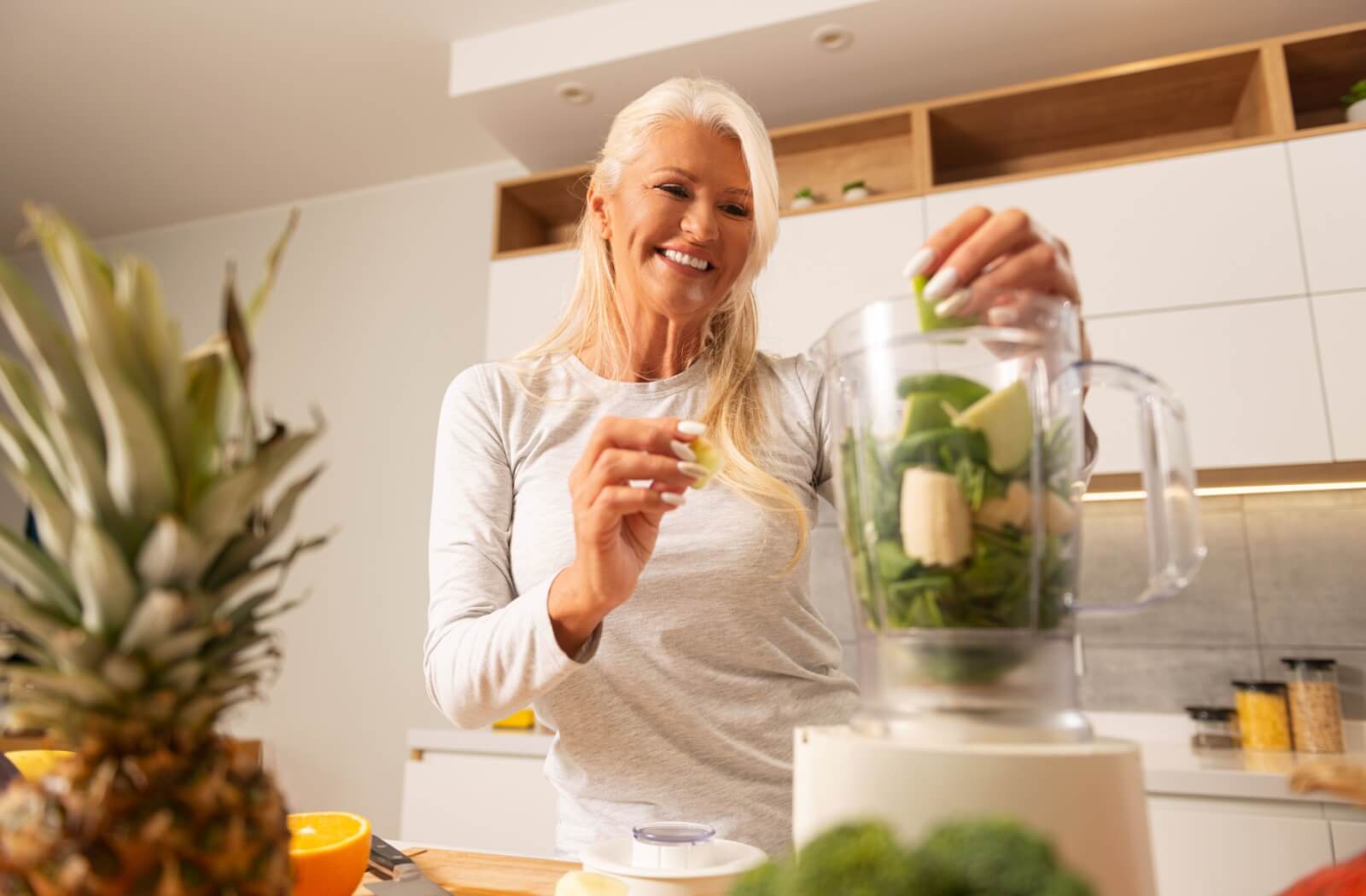 An older adult woman preparing a healthy smoothie using leafy greens and various fruits.