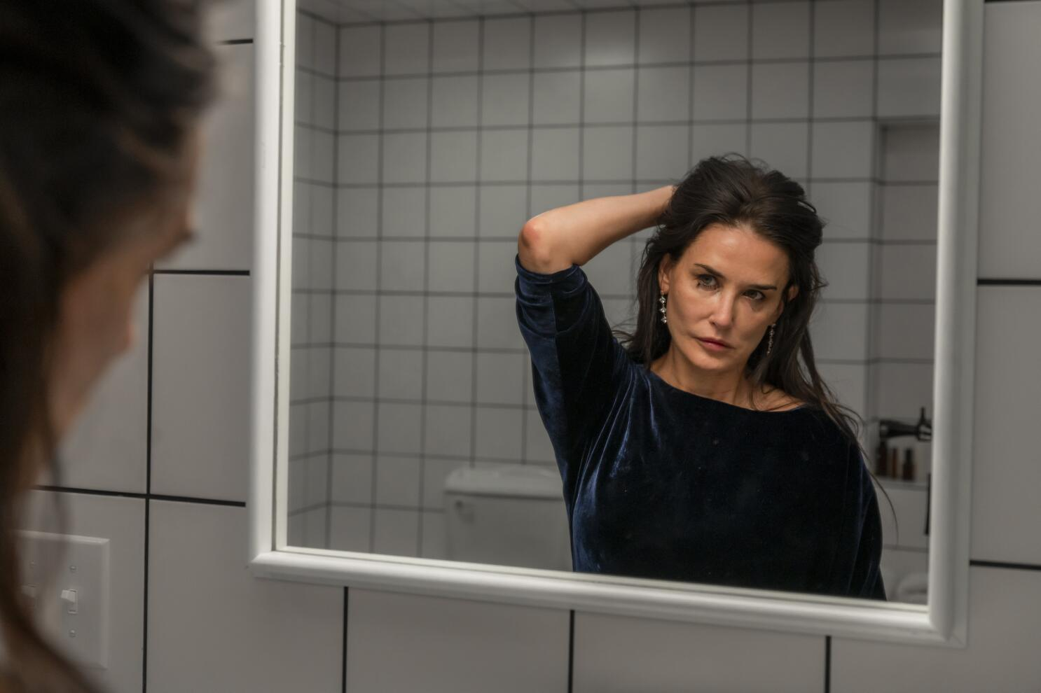 Retrato de una mujer de cabello oscuro mirando su reflejo en el espejo de un baño, con expresión seria y pensativa, sosteniendo su cabello con una mano. La pared de fondo está cubierta de azulejos blancos, creando un ambiente minimalista.