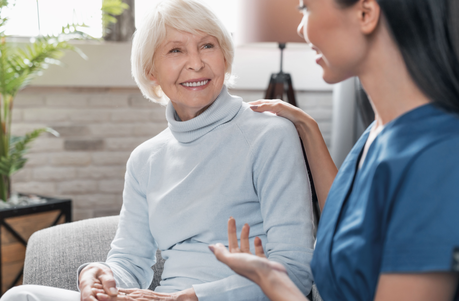 A senior resident sits with a memory care nurse chatting happily in the common area.