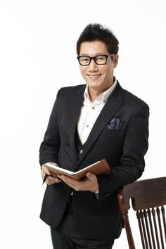 This contains an image of  Ji Suk Jin  wearing a black suit, holding a book and smiling at the camera while standing next to a chair