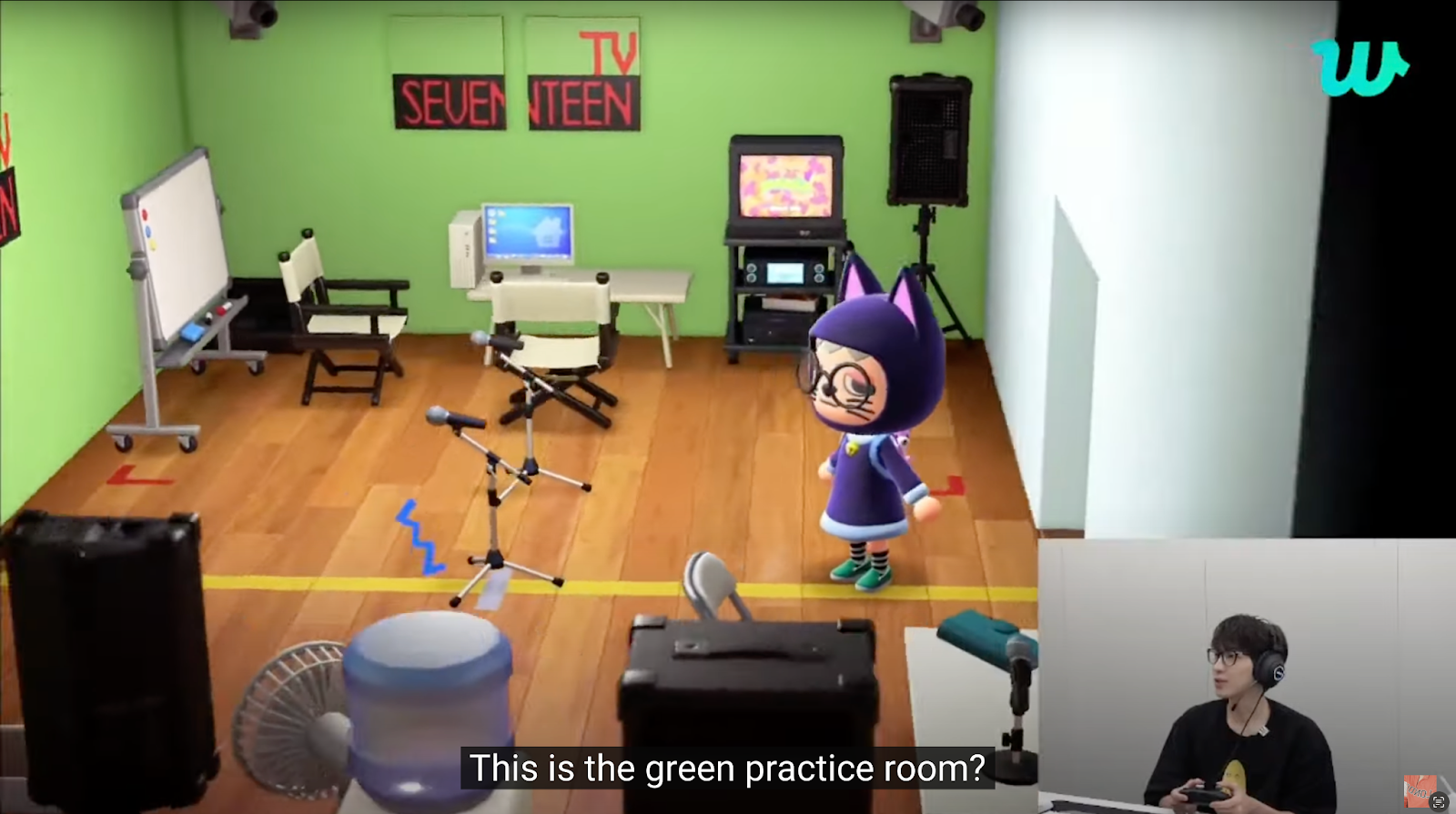 A screenshot of a livestream in which Wonwoo of Seventeen plays Animal Crossing. Here he is visiting a fan-made recreation of the ‘Melona Room’ that someone had made in Animal Crossing New Horizons. Wonwoo’s avatar is a character with glasses in a black cat costume. The avatar is standing in a room decorated to look like their old practice room, with bright  green walls, camera setups, microphones, and other equipment. The subtitle captured by the screenshot reads: “this is the green practice room?”