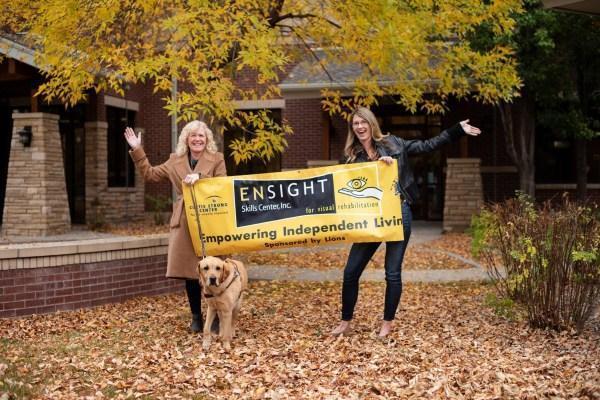 This image shows two women standing in front of a brick and concrete building with an “Ensight Skills Center” yellow banner and a golden retriever on a leash