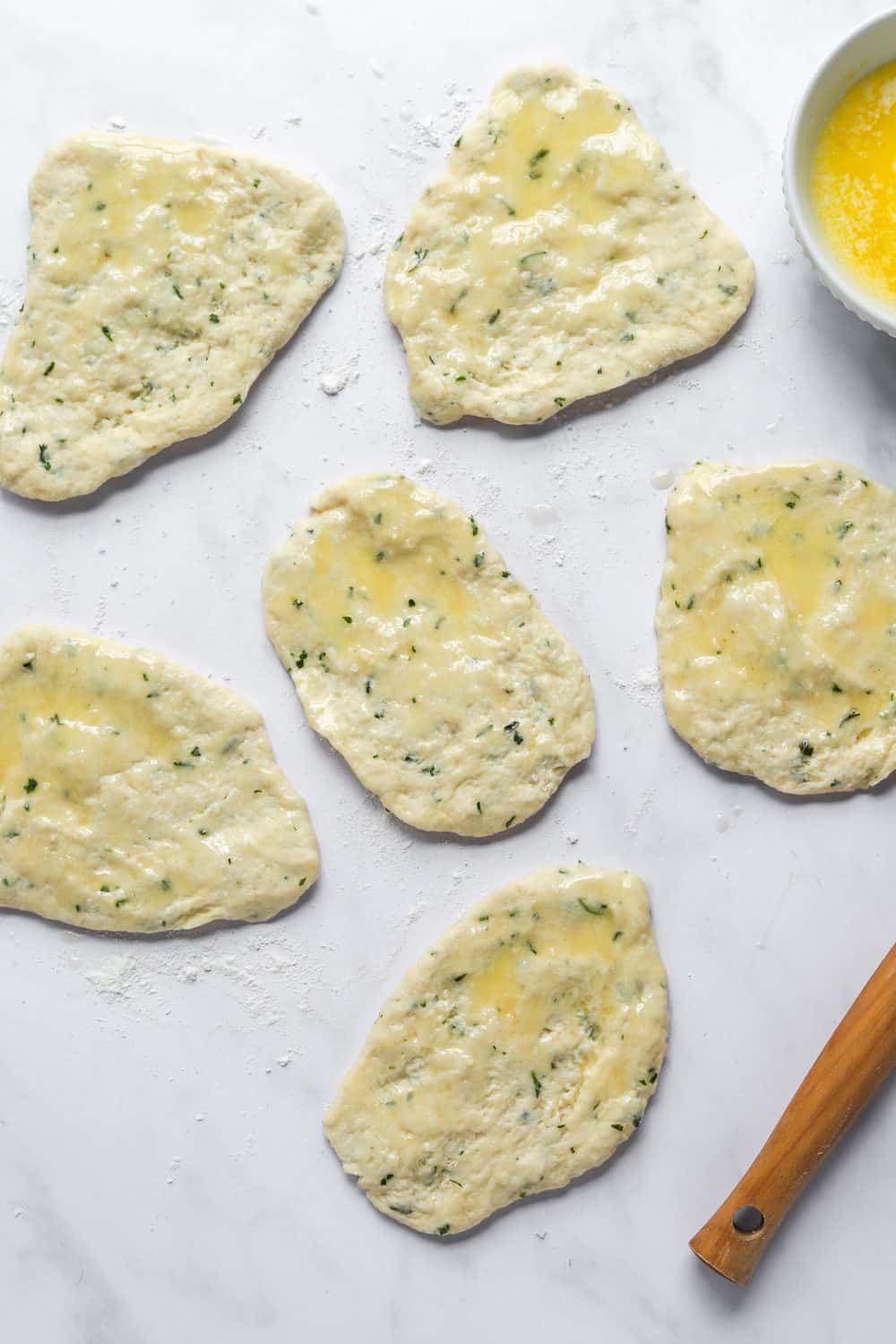 Formed uncooked naan bread dough layer out on a white surface brushed with melted butter.