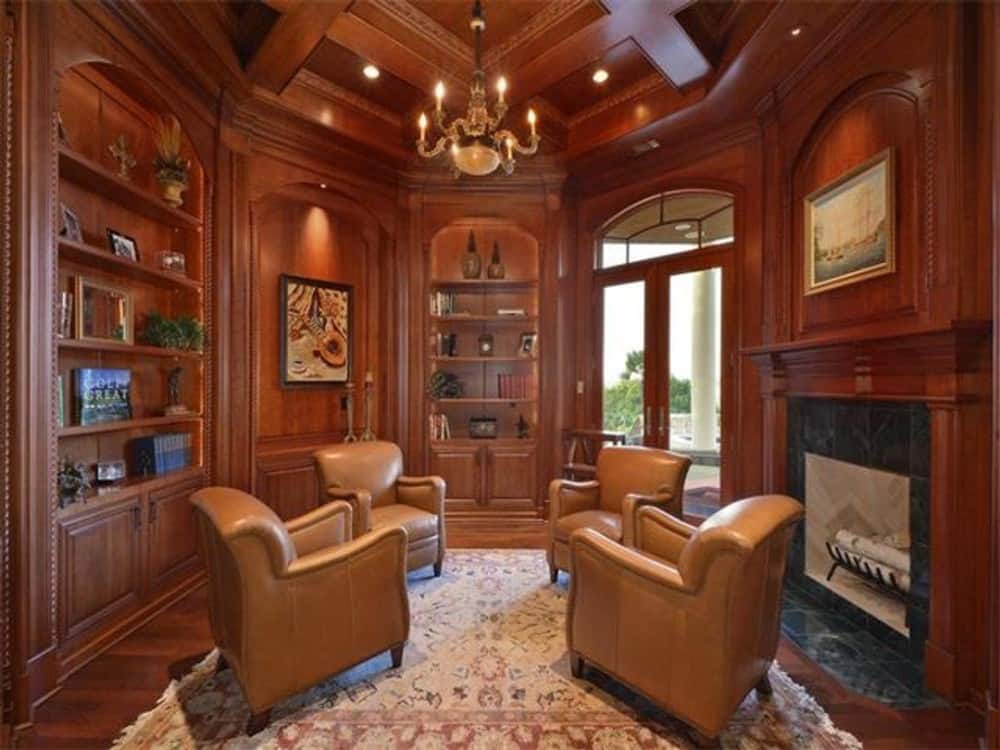 Study with leather armchairs, a fireplace, built-in shelves, and a coffered ceiling that matches the wood-paneled walls creating a cohesive look.