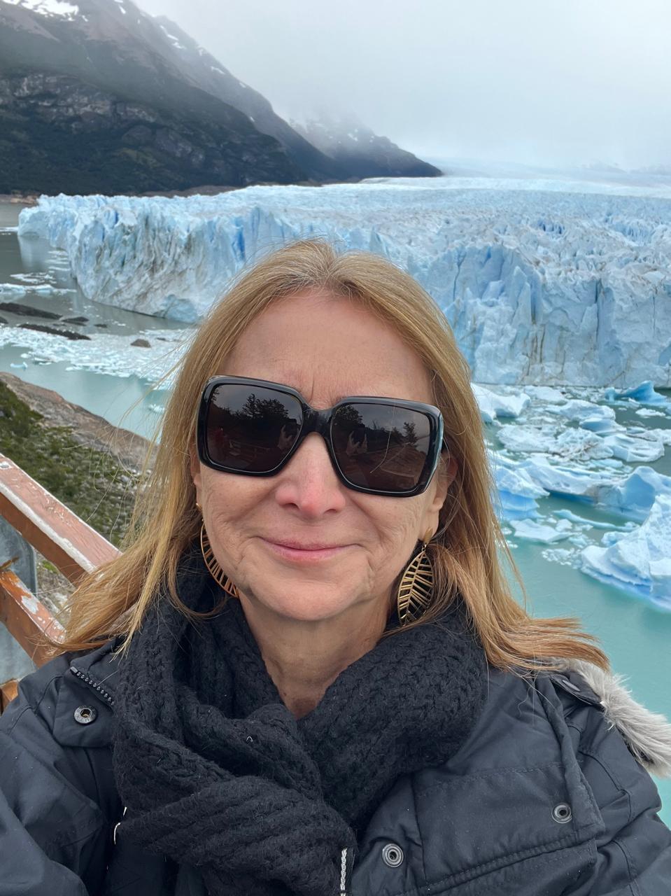 Mulher com óculos de sol na cabeça posando para foto com montanha ao fundo

Descrição gerada automaticamente