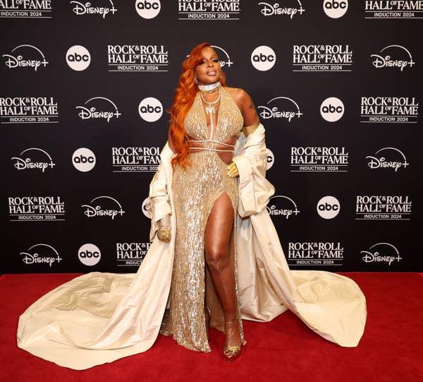 I don’t know who is in the image. The person is wearing a shimmering gown with a high slit and a long coat, standing on a red carpet with a backdrop