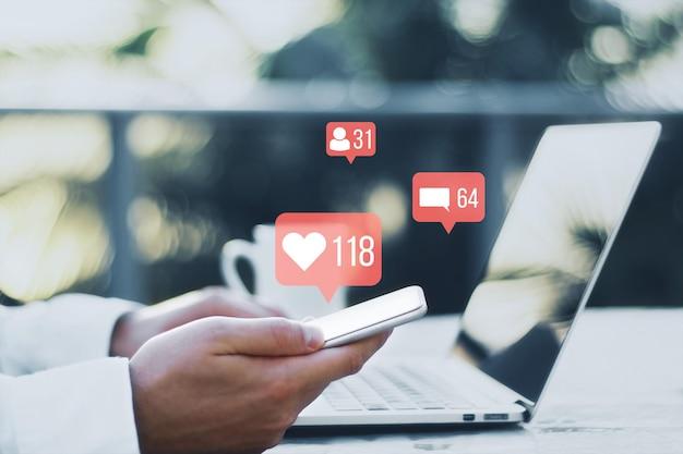 Close up of businessman hands using laptop and phone at desk with coffee cup with creative social media icons hologram on blurry outdoor background Community likes shares and communication concept