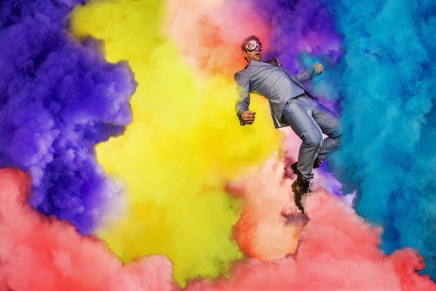 An experimental photography art piece of Chase Jarvis in a shiny silver suit, wearing Goggles, and appearing to float amongst clouds of rich purple, yellow, red, and cyan smoke.