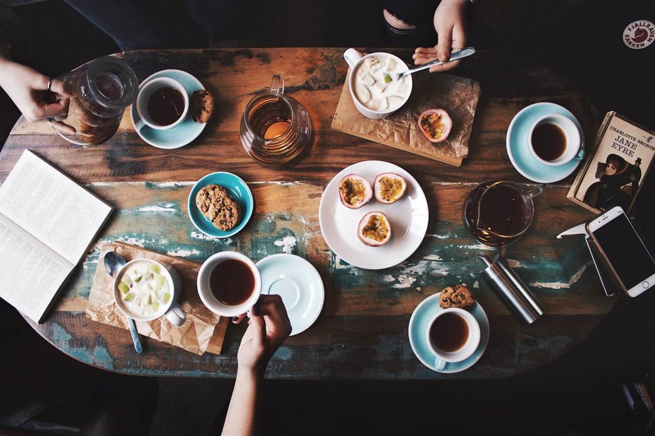 A top-down view of a table filled with coffee, tea, pastries, and fruits. The table is made of weathered wood and has a warm, inviting atmosphere. The image highlights the different ways to enjoy coffee and tea, as well as the potential for a less acidic brew.