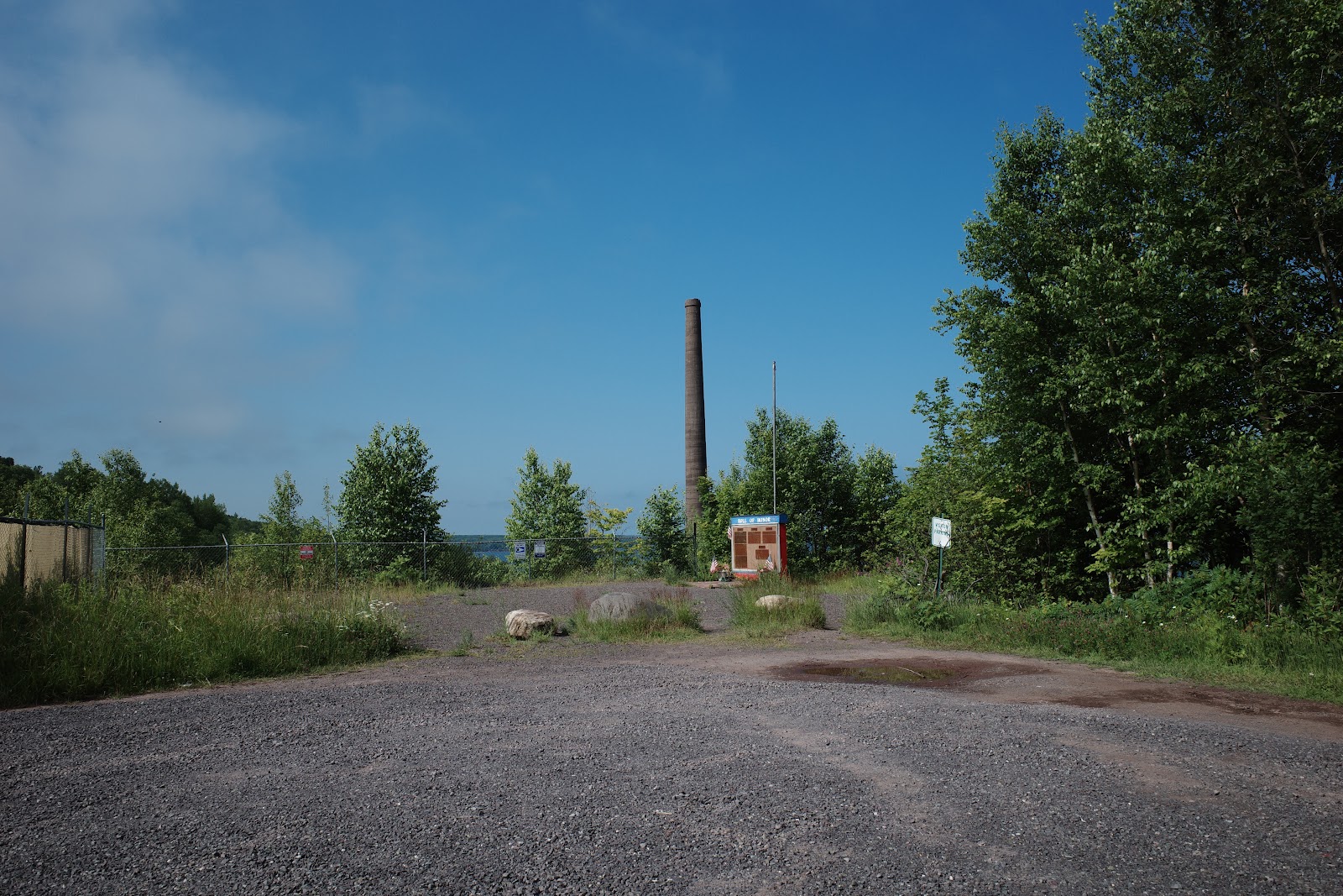 Smokestack in distance of gravel parking lot, small roadside display reading “Honor Roll” with list of names.