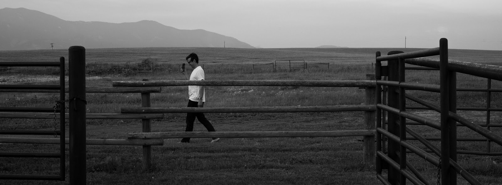 Black and white photo of a man walking across a field behind a fence.