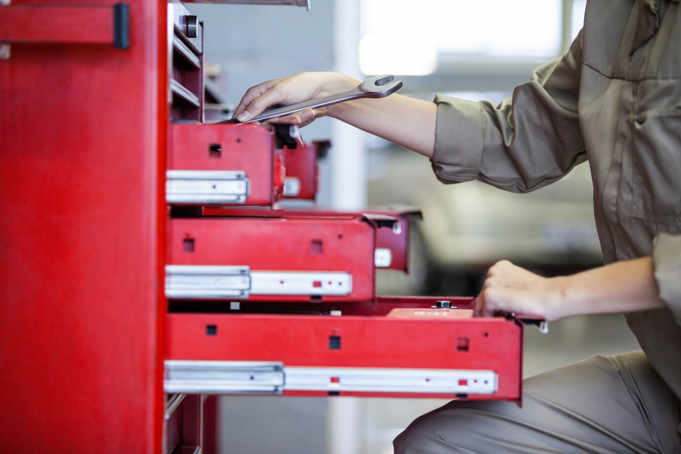 a person accessing a red tool chest with multiple drawers, commonly used in workshops or garages. The individual, dressed in a light brown work uniform, is holding a wrench while reaching into an open drawer