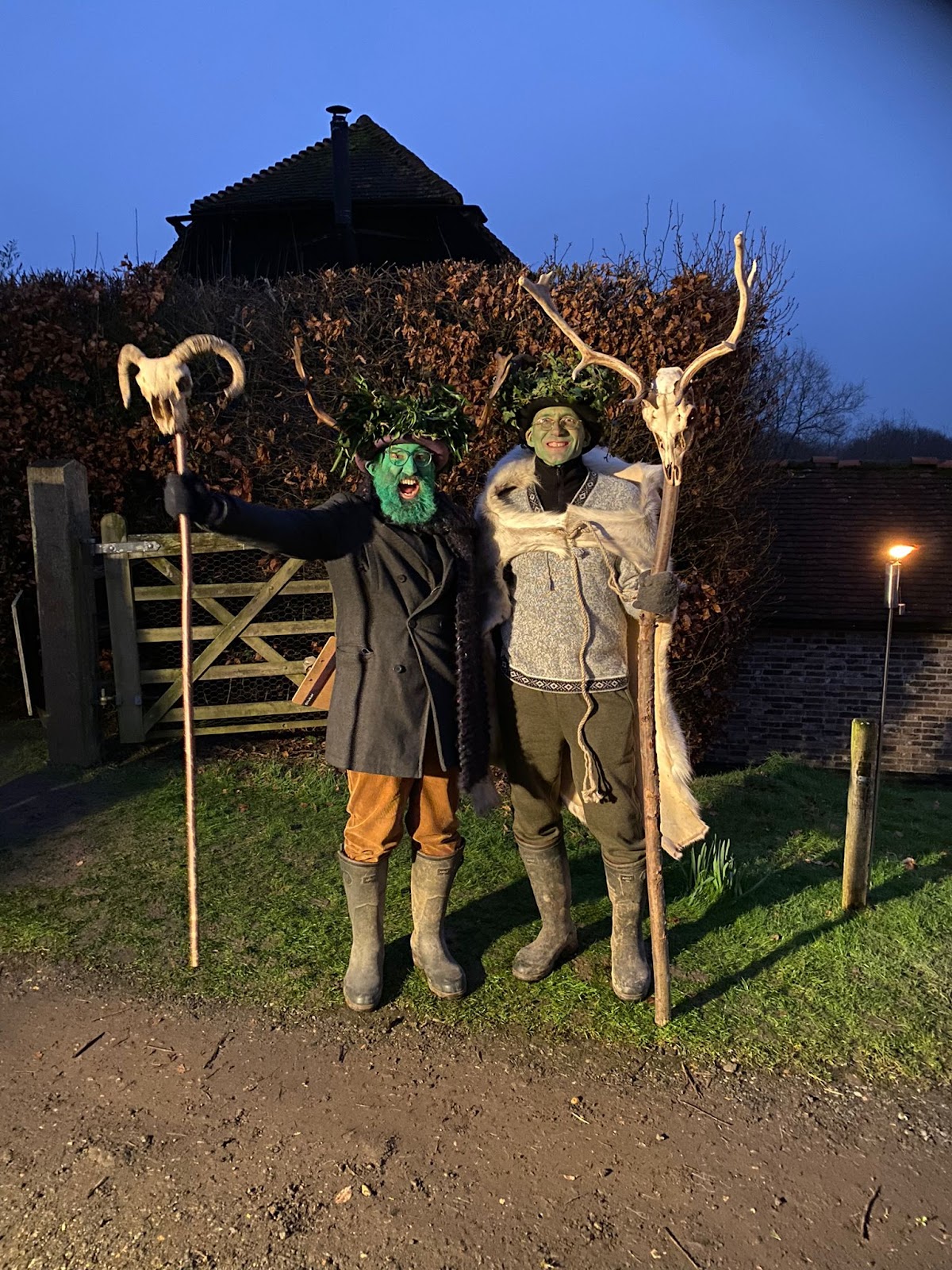 Two middle-aged white men, one with a beard and one with a big(ish) nose have their faces painted green and are wearing foliage hats. They both hold staffs with animal skulls on them.