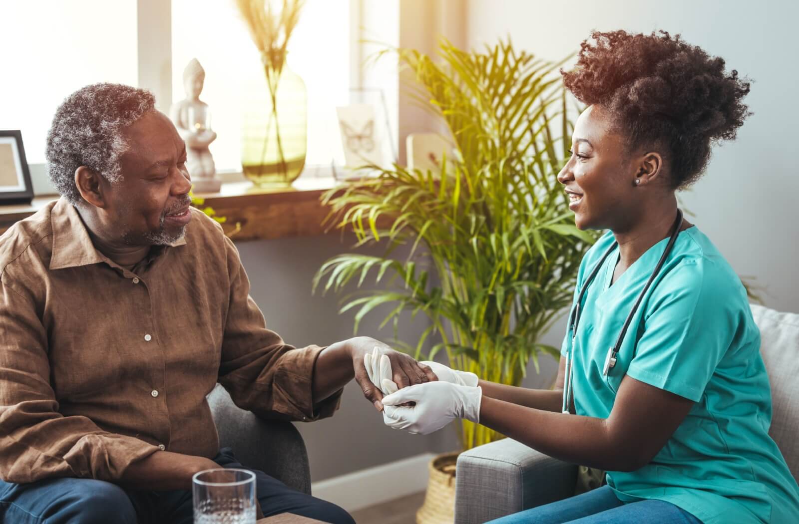 A female healthcare worker in scrubs and gloves holds the hands of an older man offering care and support.