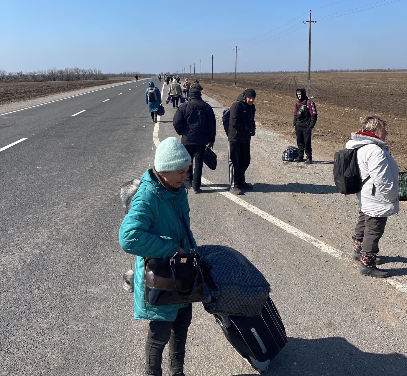 A group of people standing on a road with luggage

Description automatically generated