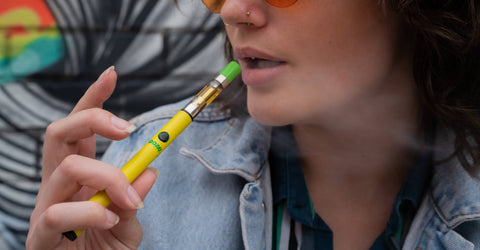 A young, white brunette woman is taking a puff from her yellow Ooze Twist Slim vape pen with an OozeX cartridge with a green tip.