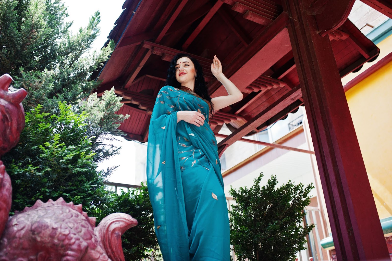 A woman in a beautiful teal saree stands under a red pavilion, exuding confidence and grace—celebrating the timeless charm of sarees on Women's Day.