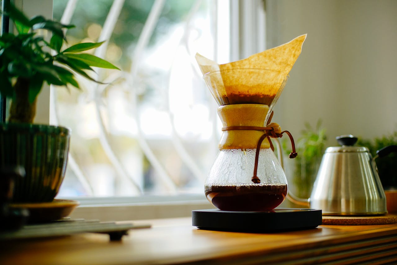 A glass Chemex coffee maker sits on a wooden counter, surrounded by a potted plant and a kettle. The coffee is brewing, and the filter paper is visible inside the cone. Sunlight streams through a nearby window, casting a warm glow on the scene.