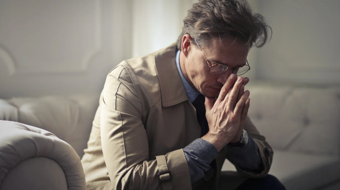 (Worried middle-aged man sitting on a couch with hands clasped in thought, conveying stress, anxiety, or legal concern in a somber setting.)