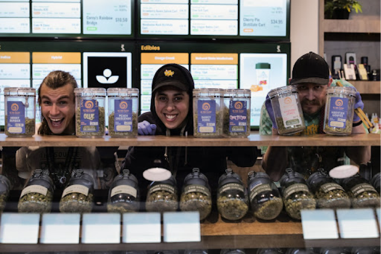 Three budtenders smile broadly while showing off The Source cannabis dispensary’s in-house-grown boutique flower, In The Flow.