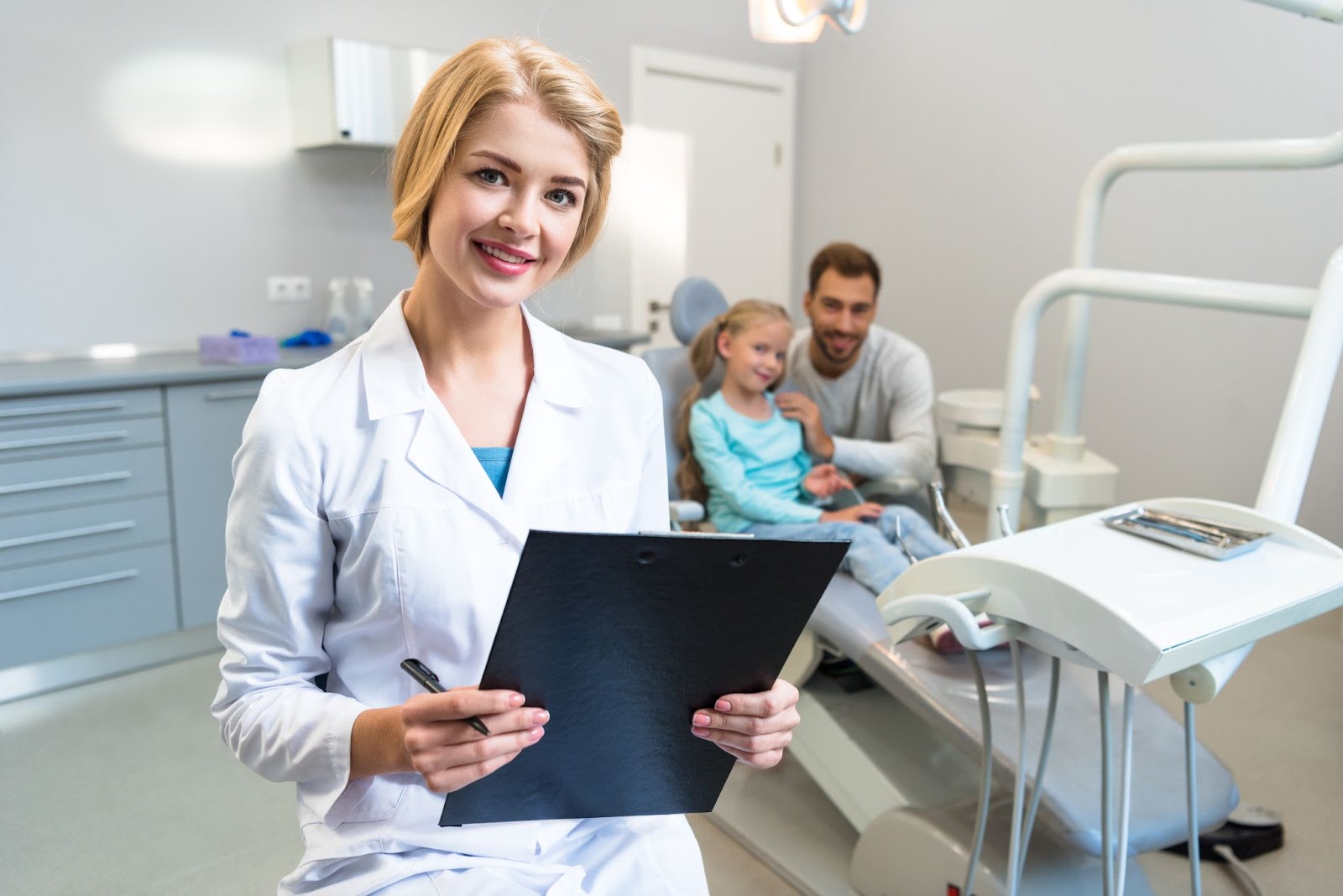 Female dentist holding a clipboard next to a man and young girl in an exam chair. 