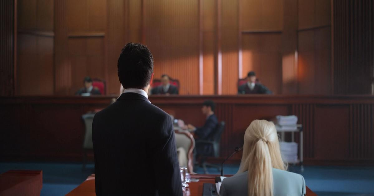 Vista de um tribunal, com foco nas costas de dois advogados ou representantes legais, um homem e uma mulher, de frente para um painel de juízes sentados ao fundo. O ambiente formal é caracterizado por mobiliário de madeira, iluminação suave e uma atmosfera típica de julgamento ou audiência.