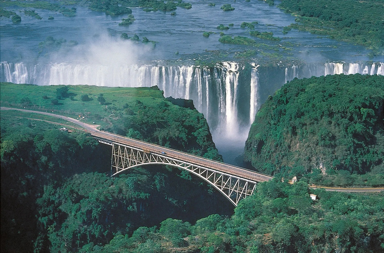 Majestic view of Victoria Falls in Zambia with a bridge crossing over the gorge, surrounded by lush greenery and cascading water from the falls, showcasing the natural beauty of the area.