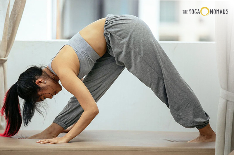 The image shows an individual performing a yoga pose on a yoga mat in a room with large windows and brick walls. The pose resembles the ‘Extended Triangle Pose’ or ‘Utthita Trikonasana,’ which is relevant for those interested in yoga practices as it demonstrates proper form and technique.