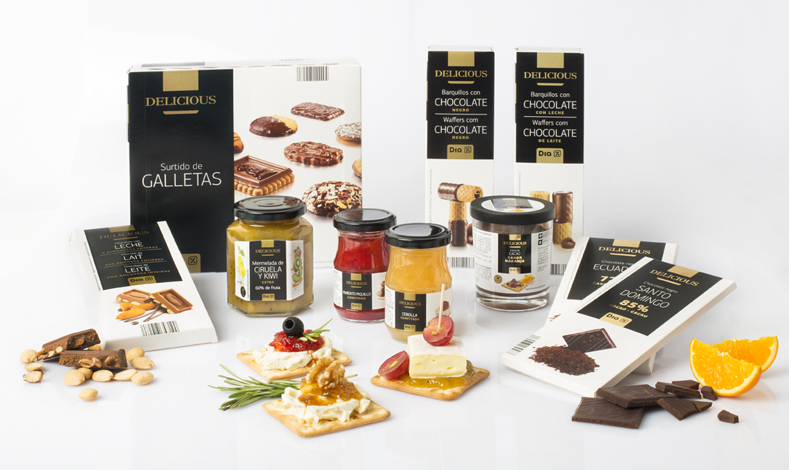 A imagem apresenta uma variedade de produtos gourmet da linha Delicious, incluindo biscoitos sortidos, barras de chocolate, wafers cobertos de chocolate, geleias de diferentes sabores, e queijos com acompanhamentos. Os produtos são dispostos de forma elegante, destacando a qualidade e a sofisticação da marca.