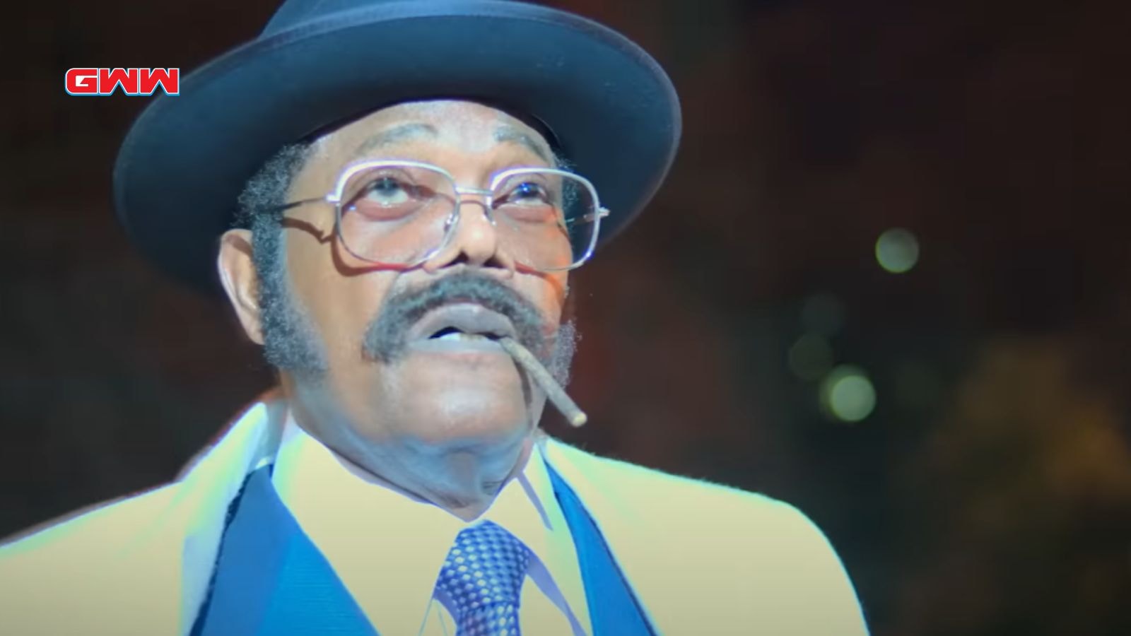 Frank Moten in hat and glasses with a cigar, illuminated at night