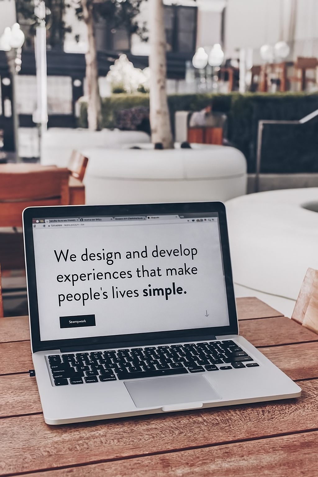 This image shows a laptop on a wooden table in a modern, outdoor cafe-style setting. The screen displays the message, "We design and develop experiences that make people's lives simple." with a button labeled "Seemywork" below it. The focus is on clarity and minimalism in digital design.