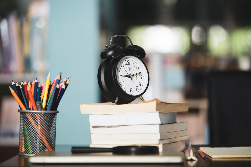 A black alarm clock sits on a stack of books on a desk, signaling it's time for a GATE Architecture 2024 crash course. The background features bookshelves, creating a blurred study environment perfect for diving into effective study tips.