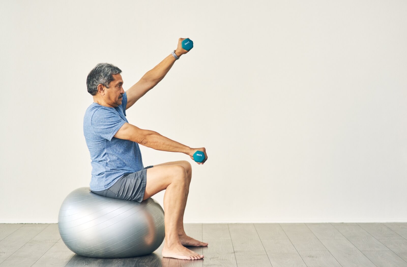 A senior man performing a balance exercise by sitting up straight on a yoga ball while lifting light weights on both hands.