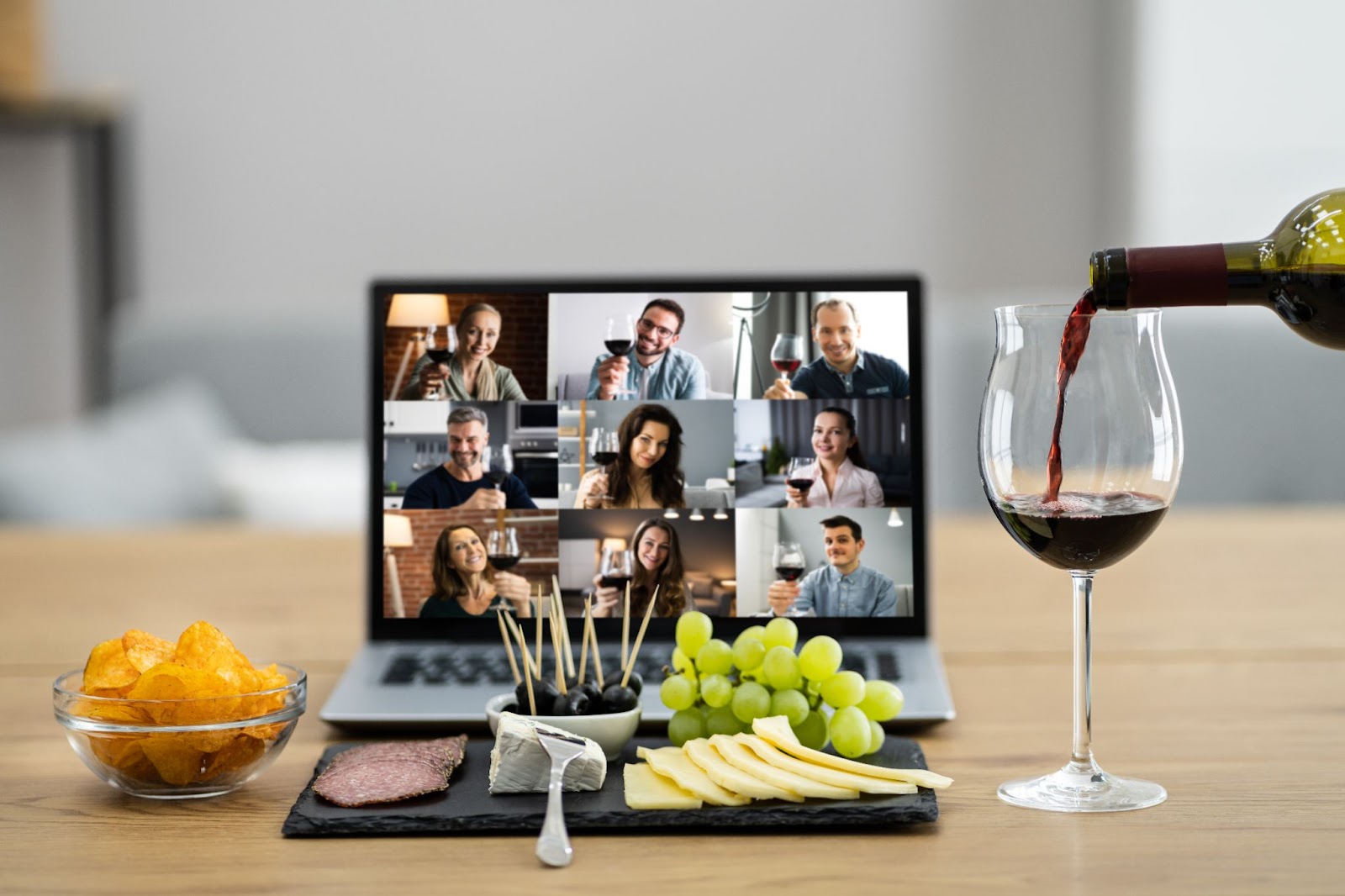 (A virtual gathering is shown on a laptop screen with nine participants. In front of the laptop, cheese, grapes, and snacks are arranged on a slate board. As a glass of wine is being poured, the group initiates the Taste and Tell Game for an evening of fun and flavor exploration.)