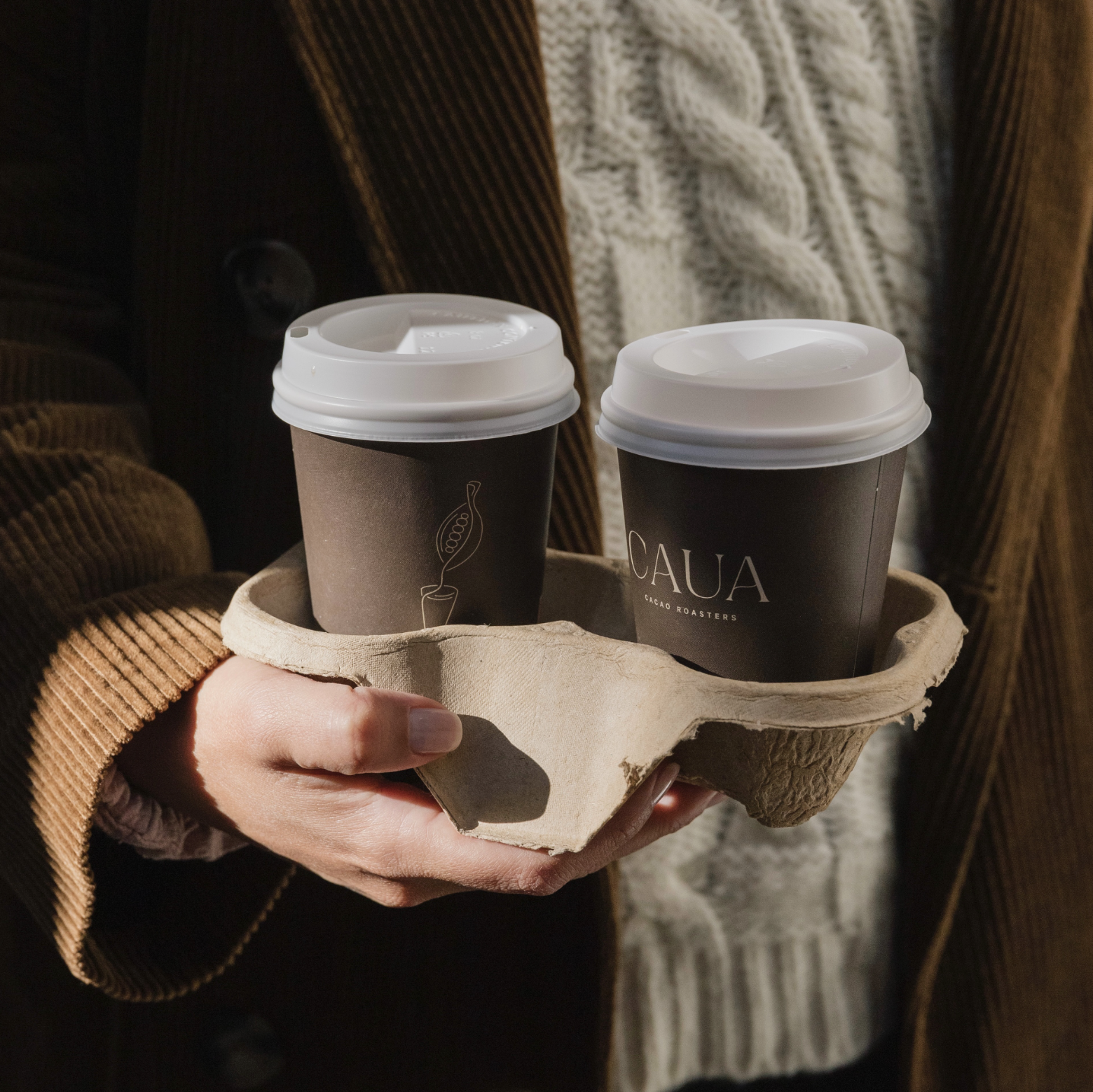 A person holds a cup holder with two dark-colored branded coffee cups.