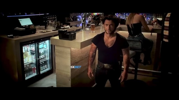 Wolverine (Hugh Jackman) chiquito en un bar. Luce una camiseta negra de escote en V y jeans con una hebilla de plata.