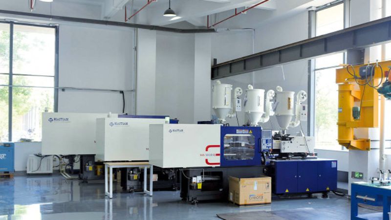 Plastic injection molding machines in a bright facility with large windows and overhead equipment.