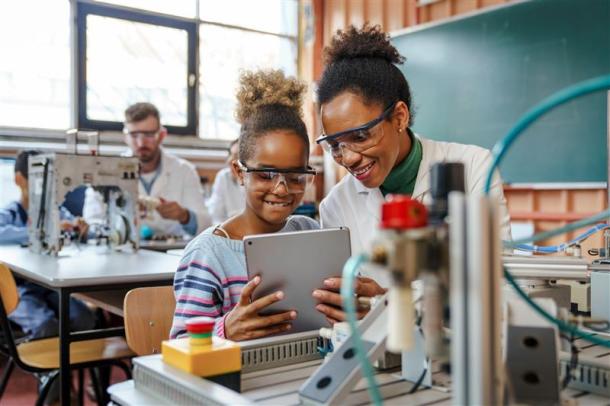 A girl uses a tablet while a female adult scientist guides her, surrounded by laboratory equipment in an educational setting.