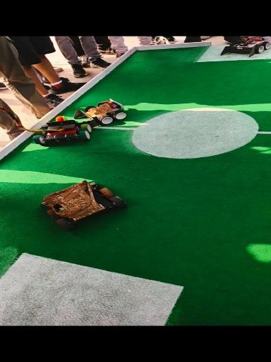 Mini robots participating in a robotics competition on a green football-themed field, with spectators gathered around.