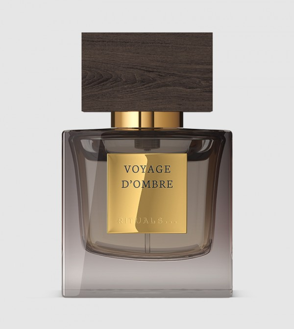 A sleek glass bottle of Voyage d'Ombre perfume by Rituals, featuring a gold label and a dark wooden cap, set against a neutral background.