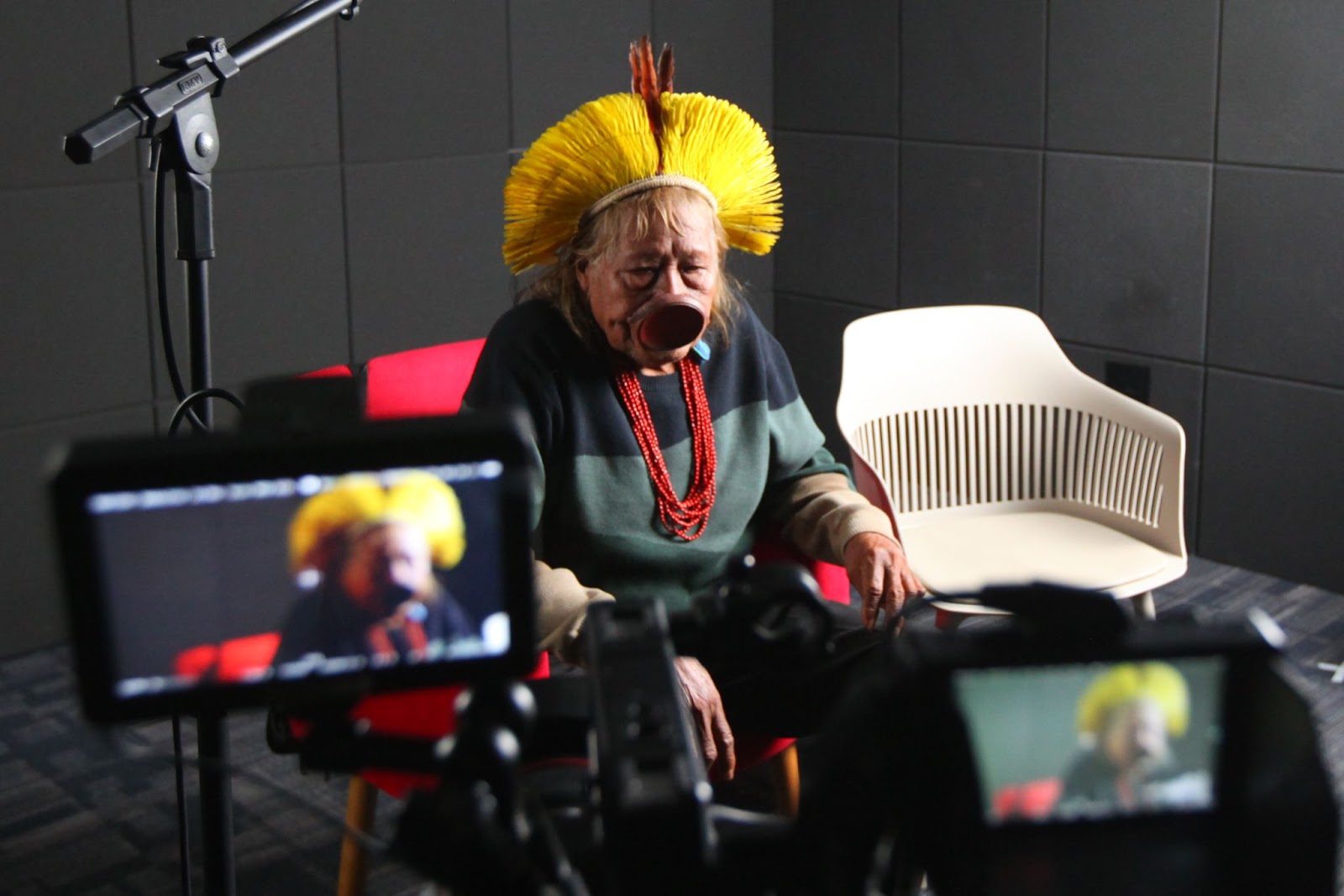 Cacique Raoni Metuktire em sua entrevista para o Museu da Pessoa. Ele está sentado em uma cadeira vermelha no estúdio. Em frente, tem uma câmera.