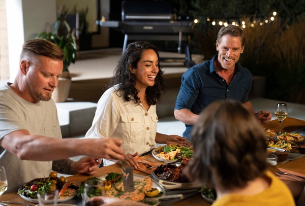 Friends gathered at night around a picnic table sharing dinner in the background a house and Traeger bbq.