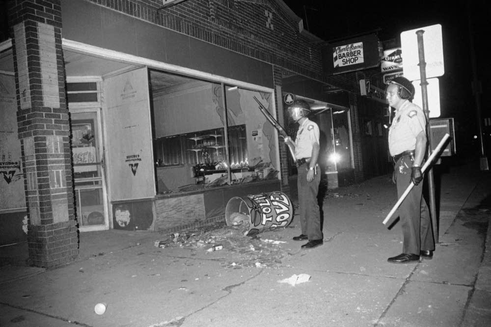 Image: Minneapolis Police during the Plymouth Ave Riots, 1967. Photo by Robert Walsh, circa 1967. A black-and-white photo showing two police officers standing in front of a store that has its front windows broken. One holds a large gun while the other holds a long baton-like rod.