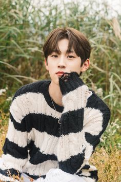 This contain DRIPPIN Minseo sitting in the grass with his hands on his face and looking at the camera