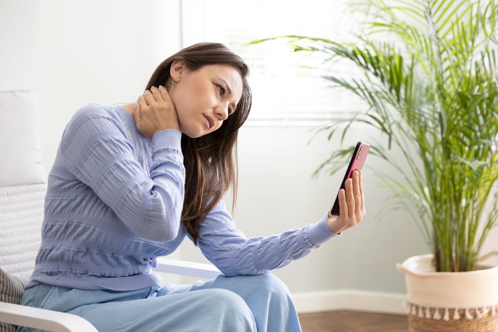 A woman with a hand on her sore neck due to bad posture while sitting and using her smartphone.