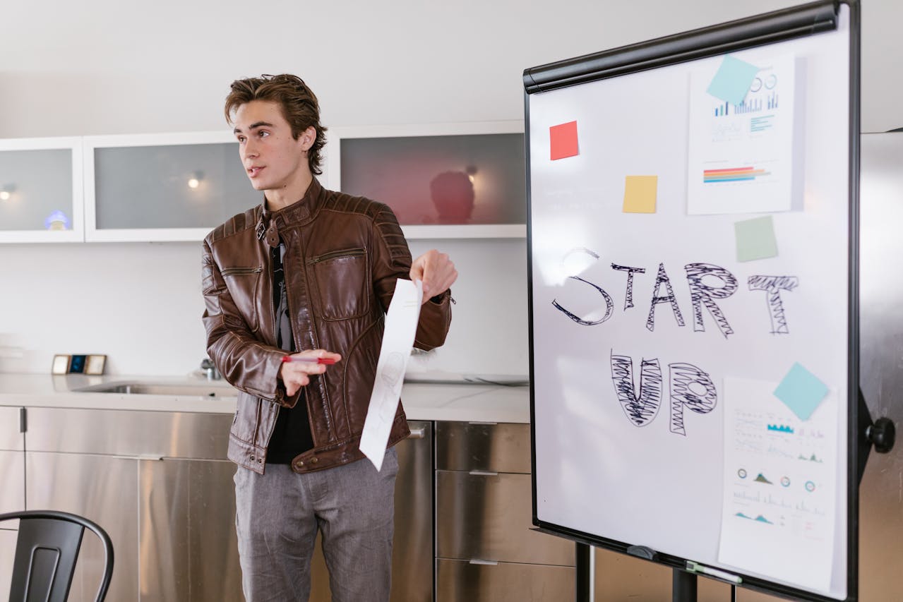 A man in a leather jacket standing in front of a whiteboard with "start up" written on it.