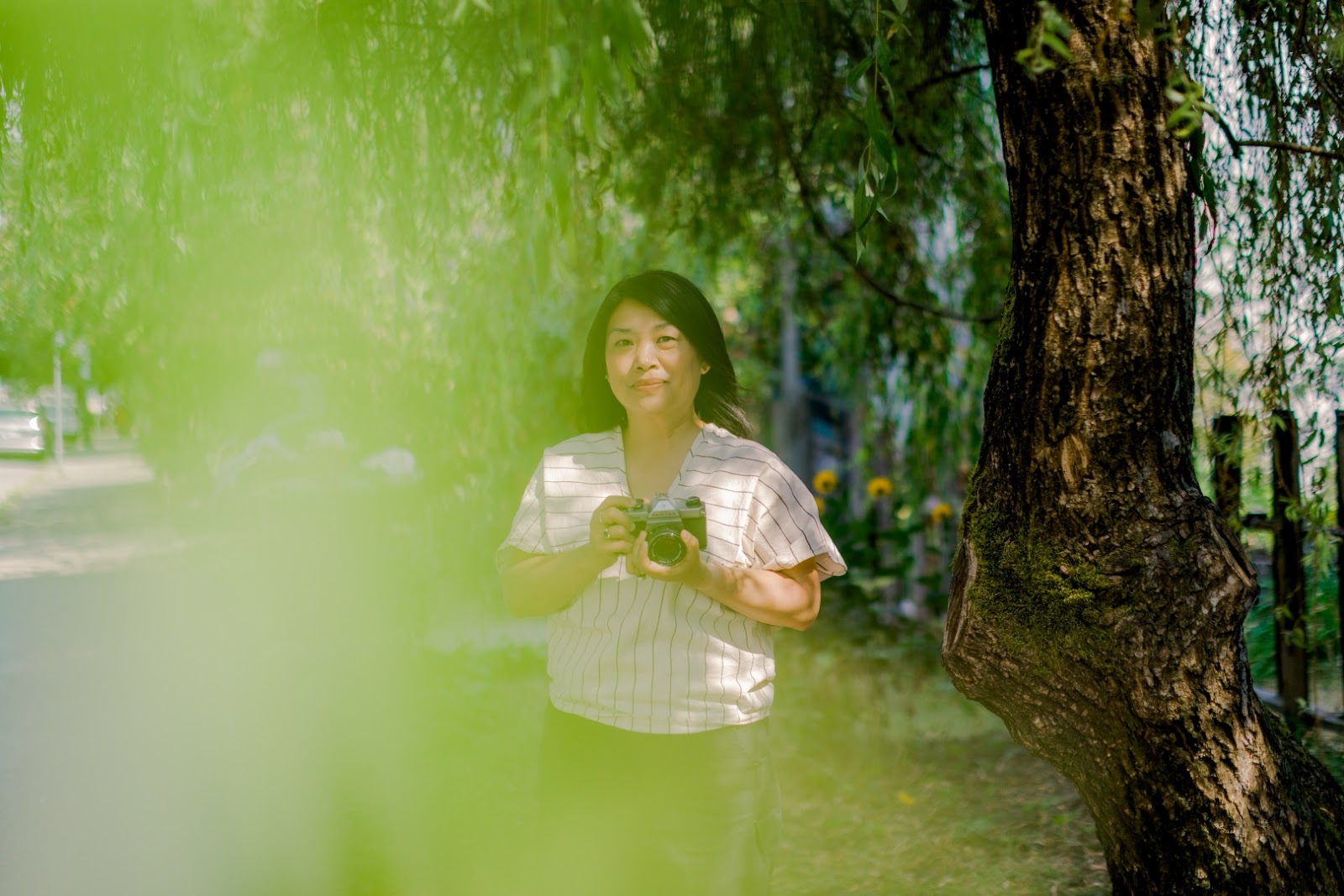 A Japanese Canadian woman holds a camera in an outdoor park, with green leaf blurring the edge of the camera