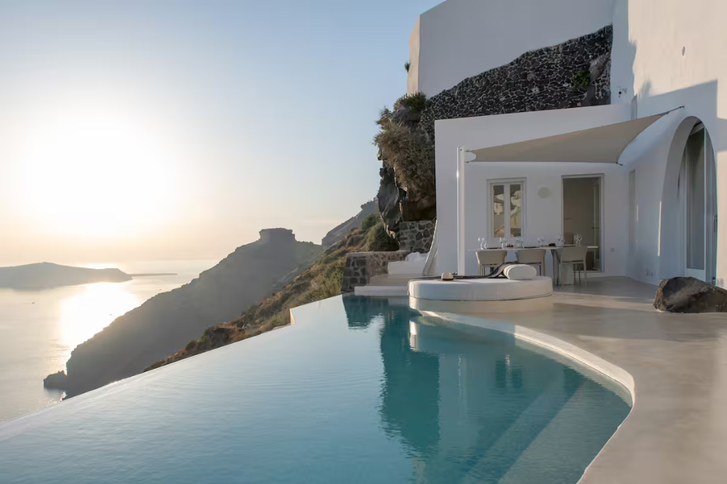 blue infinity pool looking over a cliff in santorini with a white home concrete and dining table